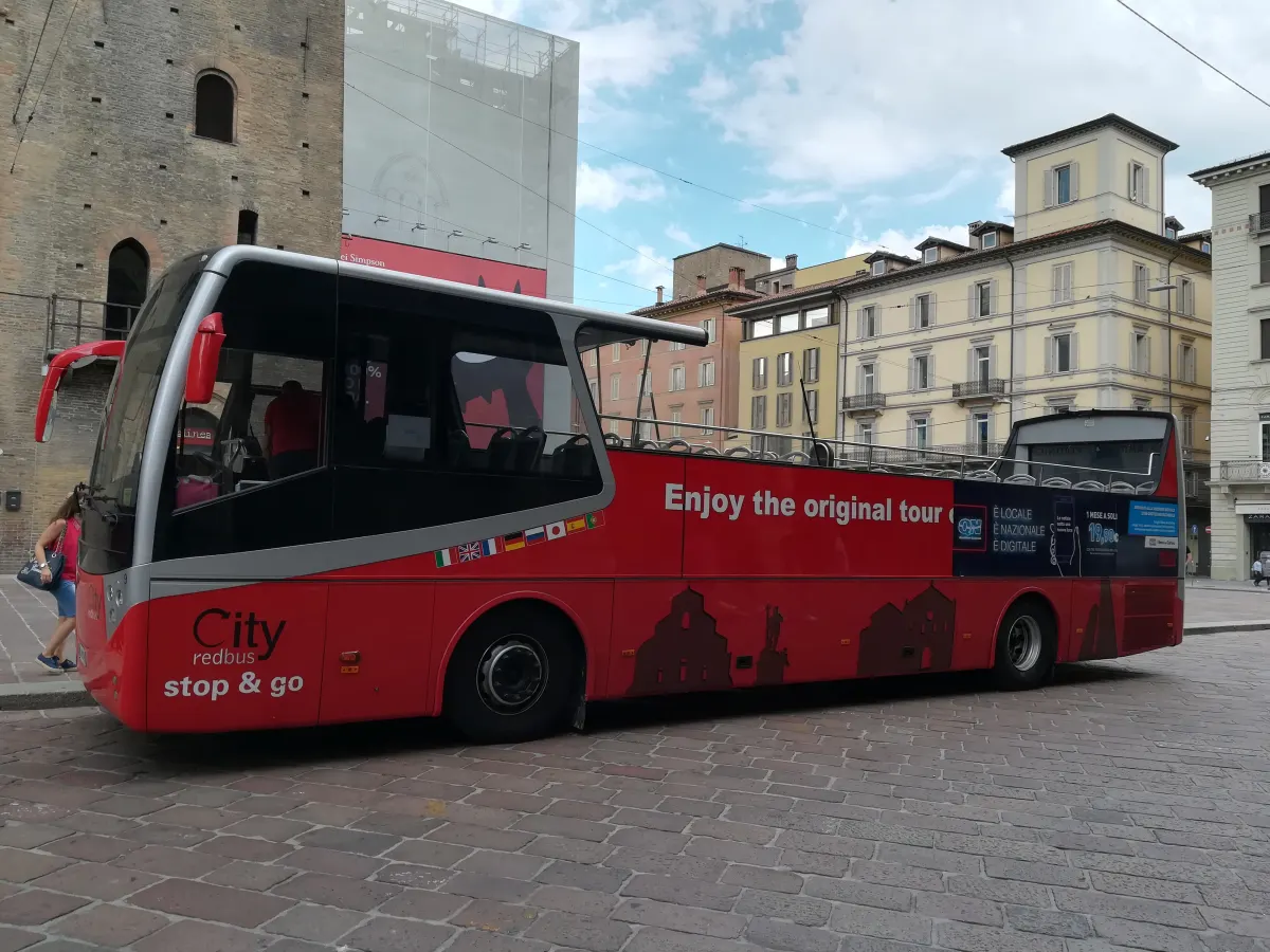 Bus touristique rouge à impériale ouverte stationné sur une place pavée de Bologne